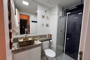 Ein Badezimmer mit einer weißen Toilette, einem Waschbecken mit Natursteinwaschbecken, einer Duschkabine aus Glas und einem Spiegel mit Kunstwerk.