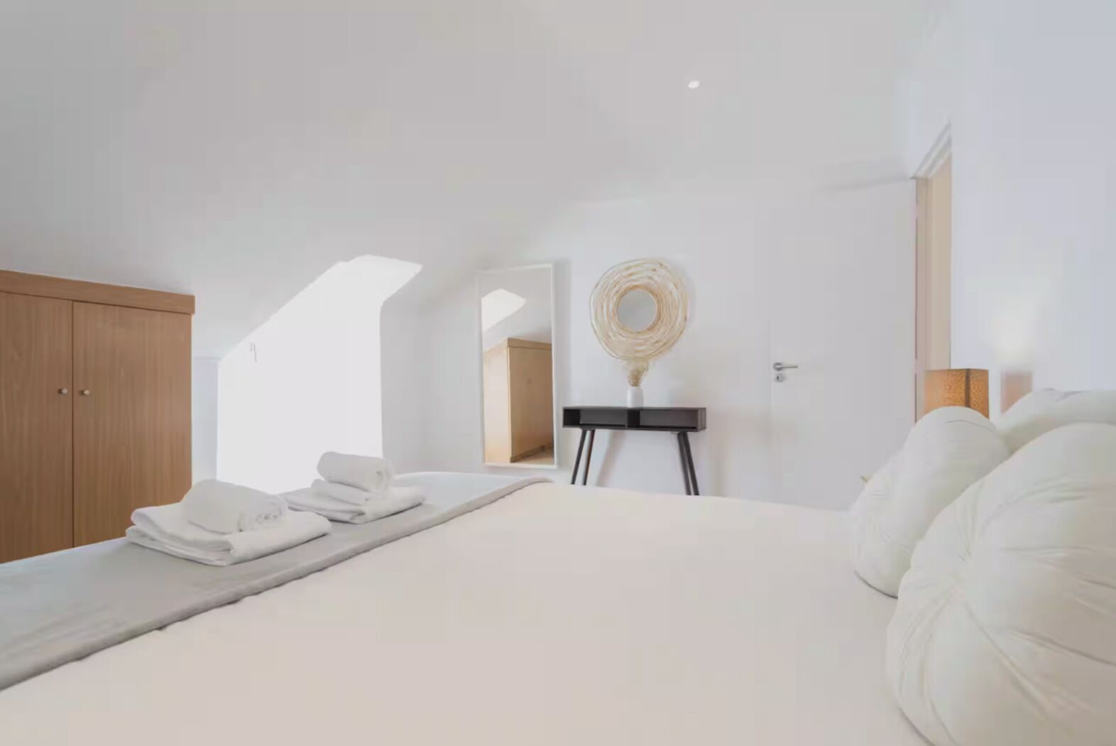 Une chambre à coucher avec un lit, une armoire en bois, une petite table sur laquelle est posé un vase, et un miroir accroché au mur.