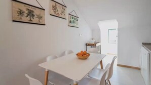 Uma sala de jantar com uma mesa e cadeiras brancas, uma tigela de laranjas e gravuras botânicas emolduradas na parede.