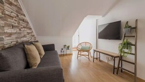 Uma sala de estar moderna com um sofá cinzento, uma poltrona de vime, uma televisão fixada na parede e piso de madeira.