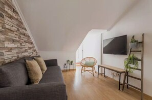 A modern living room with a grey sofa, a wicker chair, a TV mounted on the wall, and a wooden floor. - Casa Trinitária · 3BR/1WC · Sleeps 8 · Central (Lisboa)