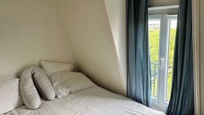 Een slaapkamer met schuine wand, een bed met witte lakens, twee kussens en een raam met gordijnen.