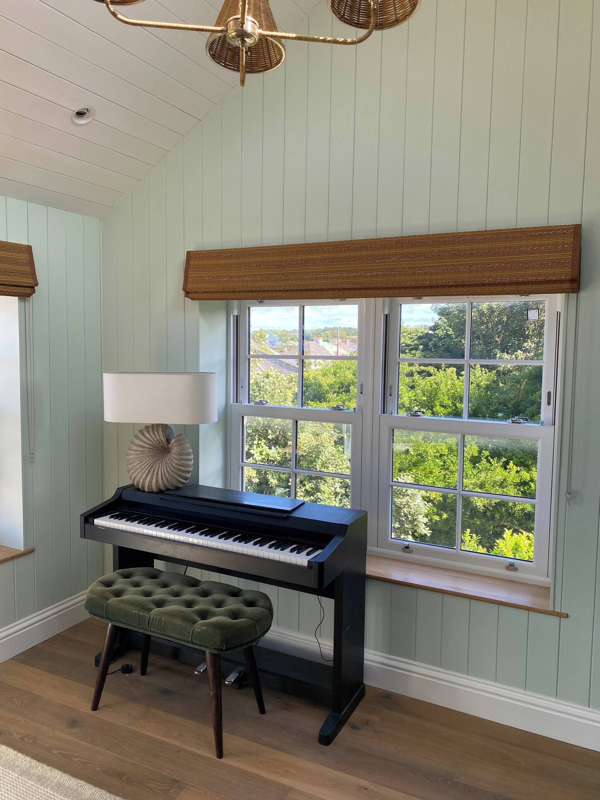 Ein Zimmer mit Klavier, einem grünen Hocker, einer Lampe und einem Fenster mit Blick auf Grünflächen.