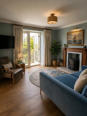 A living room with a blue sofa, a wooden floor, a fireplace, a television, and a large window with curtains. - New Stunning House St Ives With Parking (St Ives)