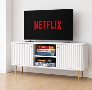 Eine weiße Unterhaltungsecke mit einem Flachbildfernseher, der 'NETFLIX' anzeigt, und drei Brettspielen: 'Catan', 'Ticket to Ride' und 'Azul'.