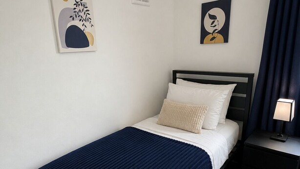 Ein Einzelbett mit einer dunkelblauen Decke, ein Nachttisch mit einer Lampe und zwei an der Wand hängende Kunstwerke.