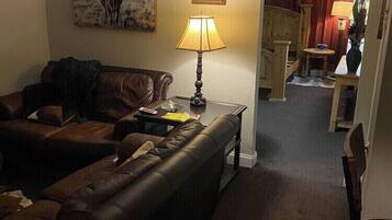 A living room with a brown leather sofa, a table with a lamp, a framed picture on the wall, and a doorway leading to another room.