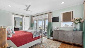 Une chambre à coucher avec un lit, un ventilateur de plafond, une télévision, une fenêtre avec des rideaux et une vue sur l’extérieur.