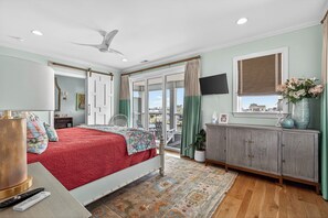 Ein Schlafzimmer mit Bett, Deckenventilator, Fernseher, Fenster mit Vorhängen und Blick nach draußen.