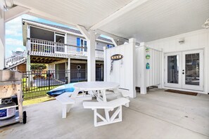 Ein überdachter Patio-Bereich mit weißem Tisch und Bänken, einem Grill und Blick auf ein zweistöckiges Gebäude mit Balkonen.