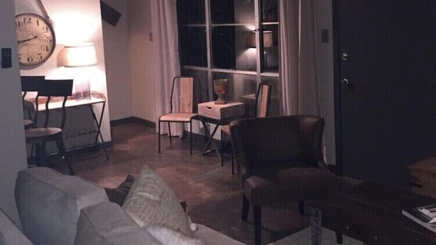 Un salon avec un canapé, des fauteuils, une table et une lampe. Il y a une fenêtre avec des rideaux et une porte.