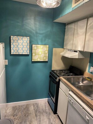 A compact kitchen with teal walls, a gas stove, a dishwasher, and two patterned wall art pieces. - 2720 #104 Oaklawn furnished rental (Dallas)