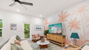 Un salón con un sofá, una mesa de centro, un televisor sobre un mueble de madera y una pared con estampado de palmeras.