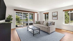 Una sala de estar con un sofá, una mesa de centro de vidrio y un televisor. Hay ventanas amplias y vistas a un edificio.