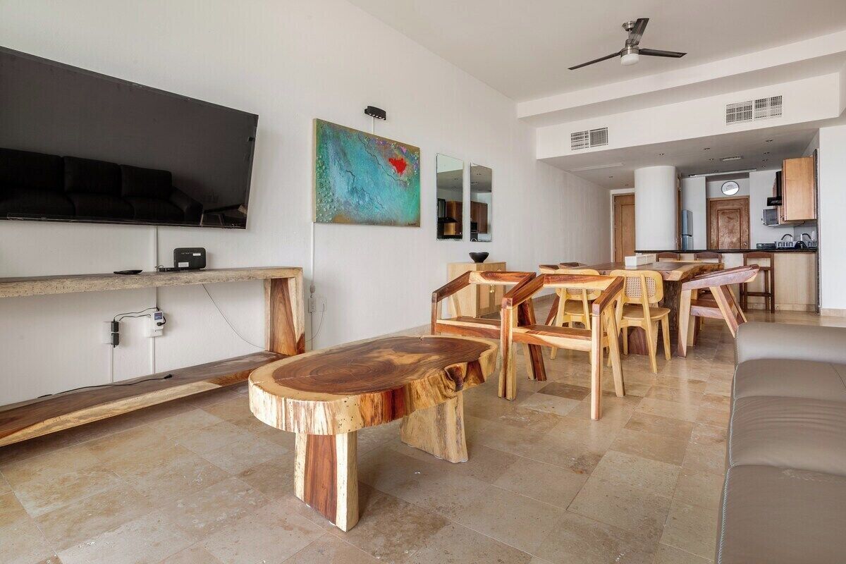 Uma sala de estar moderna com uma televisão de tela plana, móveis de madeira e uma cozinha ao fundo.