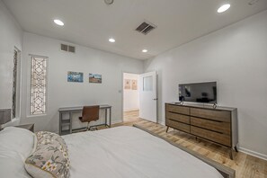 Un dormitorio con cama, escritorio, cómoda, televisor y puerta que da a otra habitación.