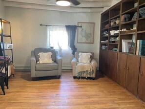 A living room with two armchairs, a wooden bookshelf, and a window with curtains. - Classic Historic Adobe Home on Kit Carson Road in Taos (Taos)