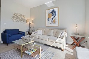 A living room with a blue sofa, a white sofa, a glass coffee table, a floor lamp, and a painting on the wall.