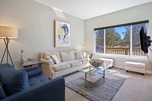 A modern living room with a sofa, coffee table, and a large window with a view of trees. - Present Pheasant - Cute & Updated, Close to Everything, Golf, Amenities, Perfect For Family (Redmond)