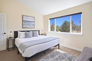 A bedroom with a bed, a nightstand, a lamp, a window with a view of trees, and a painting on the wall.