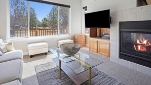 A modern living room with a fireplace, a television, and a large window overlooking a deck. - Present Pheasant - Cute & Updated, Close to Everything, Golf, Amenities, Perfect For Family (Redmond)