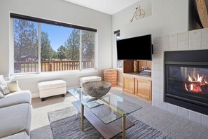 A modern living room with a fireplace, a television, and a large window overlooking a deck. - Present Pheasant - Cute & Updated, Close to Everything, Golf, Amenities, Perfect For Family (Redmond)