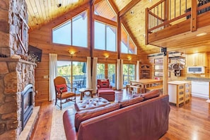 Un intérieur spacieux de chalet en bois, avec une cheminée, un canapé en cuir, un coin repas et de grandes fenêtres.