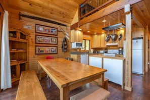 Interior de una cabaña de madera con comedor, zona de cocina y habitación en el altillo.