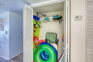A storage closet with shelves and a hanging rack, containing various items like a green inflatable ring, a blue and green inflatable ring, and a green bag. - King's Oasis Oceanview Pool BBQ Grill (Daytona Beach)