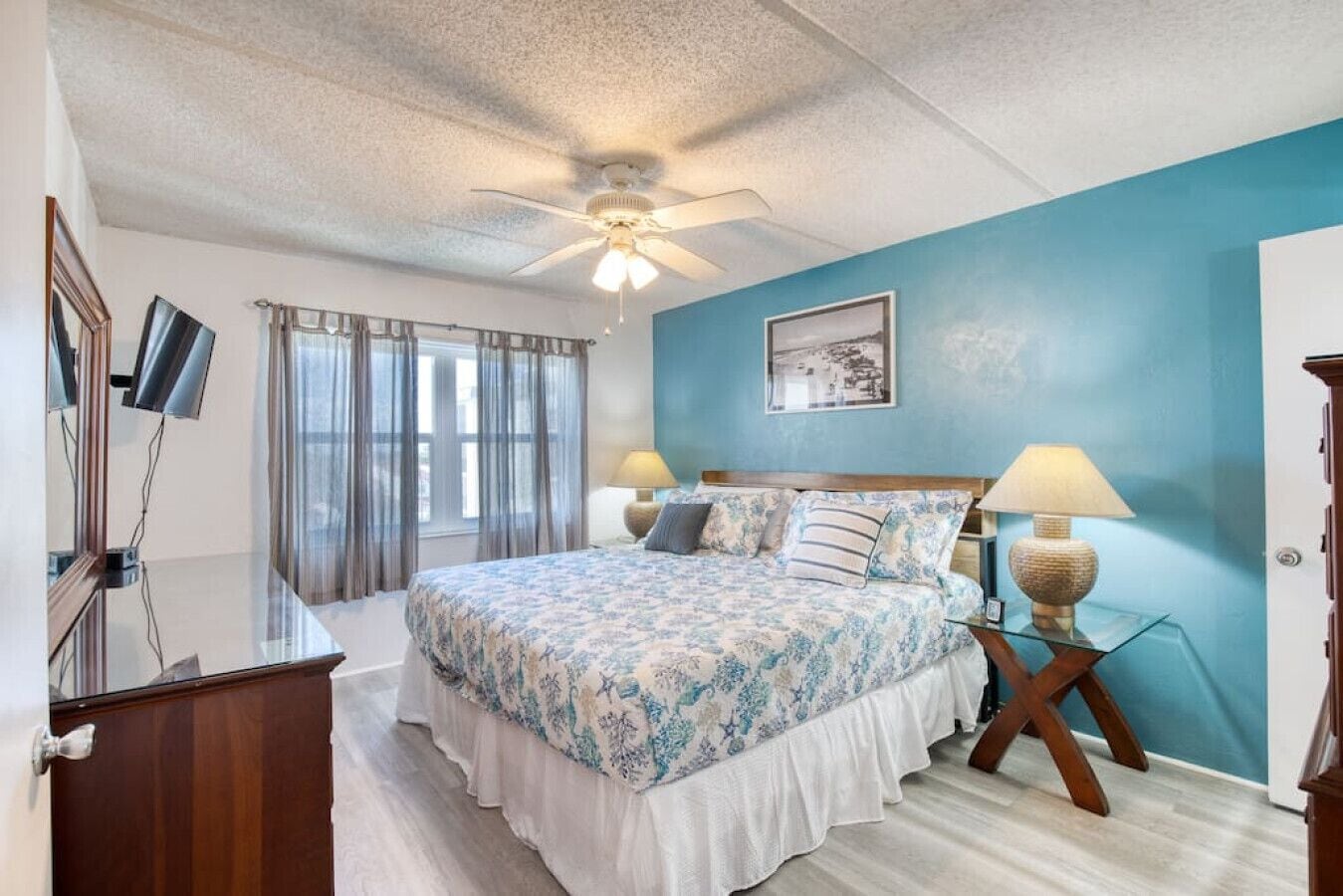 A bedroom with a bed, two bedside lamps, a ceiling fan, a window with curtains, and a wall-mounted TV.