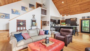 A spacious living area with a sofa, ottoman, and patterned chairs, a kitchen with dark cabinets, and a wooden ceiling. - Beautiful and comfy mountain view home with outdoor living showcased! (Waynesville)
