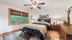 Ein Schlafzimmer mit Bett, Deckenventilator, Fenster mit Jalousien, Nachttisch, Kommode und Teppich.