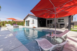 A swimming pool with lounge chairs and a red umbrella, a barbecue area, and a house with a tiled roof. - Zuri Eight - Modern 3BR Oasis w Pool (Indio)