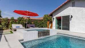 A swimming pool with a waterfall feature, a red umbrella, and a lounge area with white and blue cushions. - Zuri Eight - Modern 3BR Oasis w Pool (Indio)