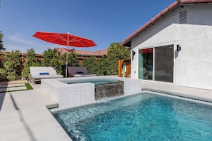 A swimming pool with a waterfall feature, a red umbrella, and a lounge area with white and blue cushions. - Zuri Eight - Modern 3BR Oasis w Pool (Indio)