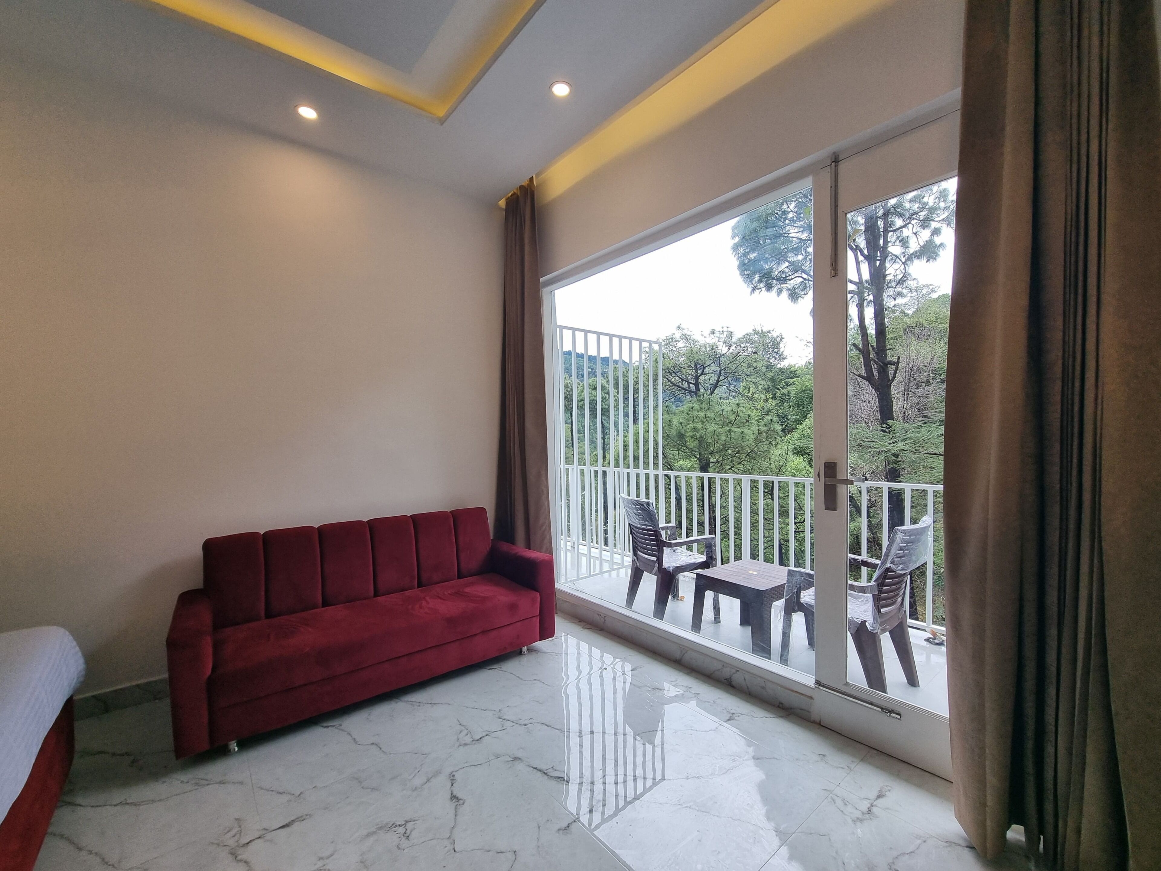 Una habitación con un sofá rojo, una cama, un balcón con mesa y sillas, y vistas a árboles.