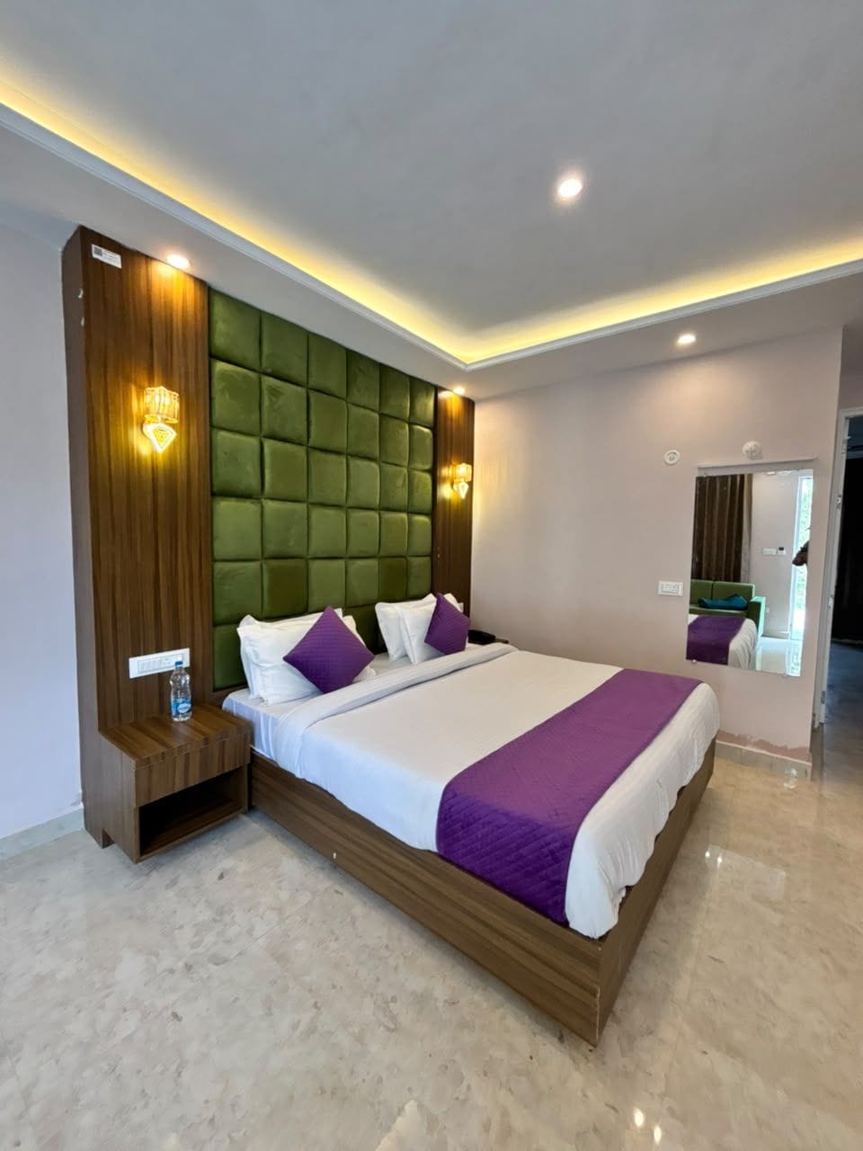 Una habitación de hotel moderna con una cama grande, una mesita de noche de madera, un cabecero acolchado verde y un espejo en la pared.