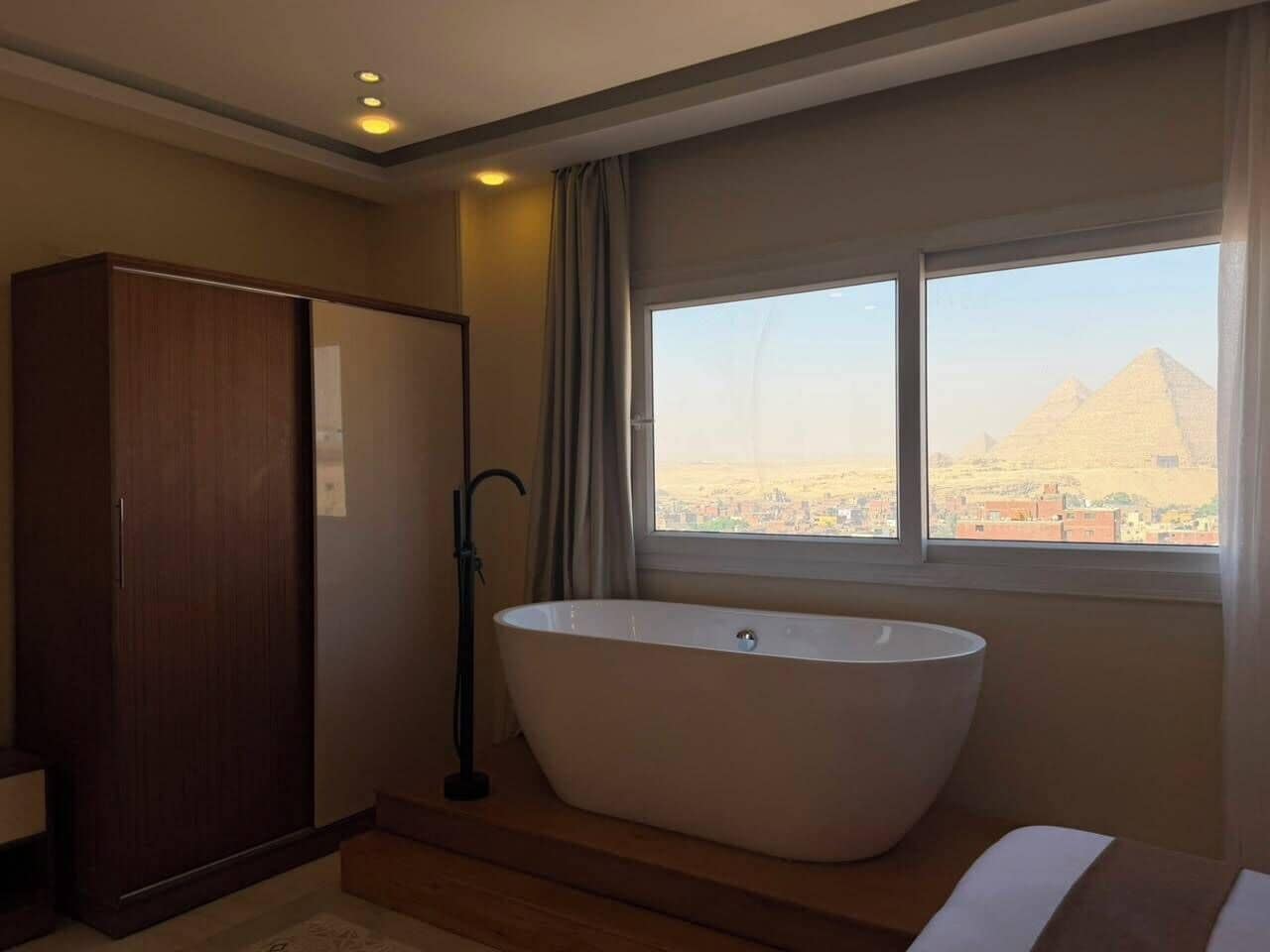Un baño moderno con una bañera independiente, un armario y una ventana con vistas a las pirámides.