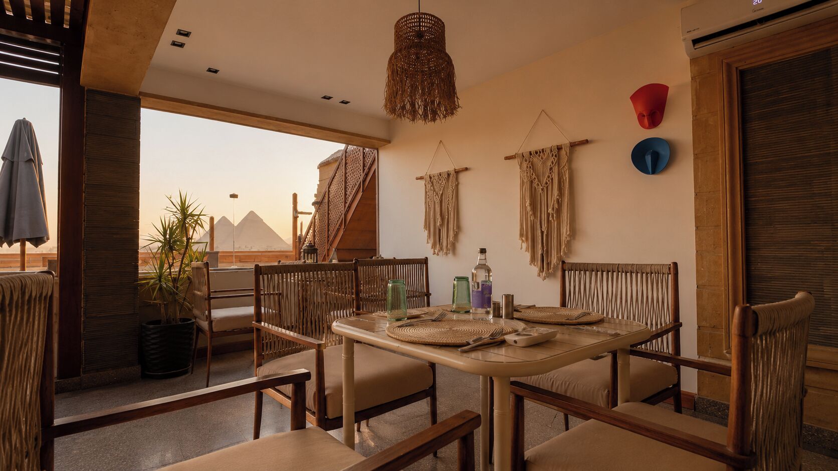 Un comedor con una mesa lista para dos personas, rodeado de sillas de mimbre, y una vista de pirámides al fondo.