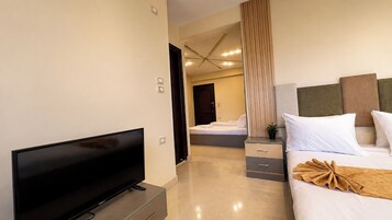 Habitación de hotel con televisión de pantalla plana, cama con ropa blanca, mesita de noche con planta y cabecera moderna.