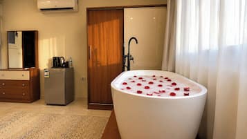 Un baño con una bañera independiente llena de pétalos de rosa, una puerta de madera, un espejo, una cafetera y un refrigerador.