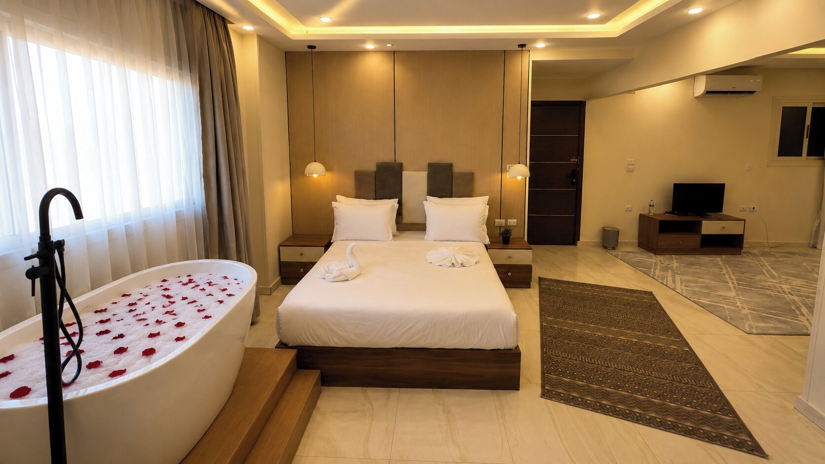 Habitación de hotel con una cama grande, una bañera con pétalos de rosa, un televisor de pantalla plana y un escritorio con una silla.