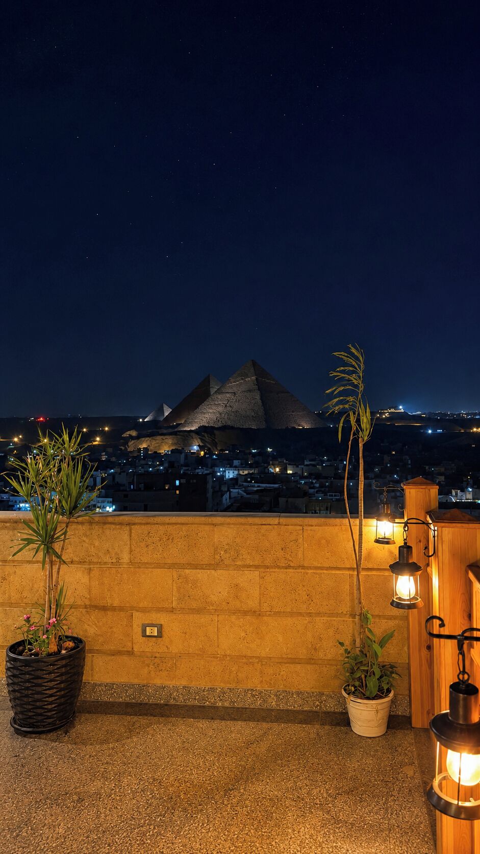 A rooftop terrace with a view of a pyramid-shaped structure, potted plants, and a cityscape at night.