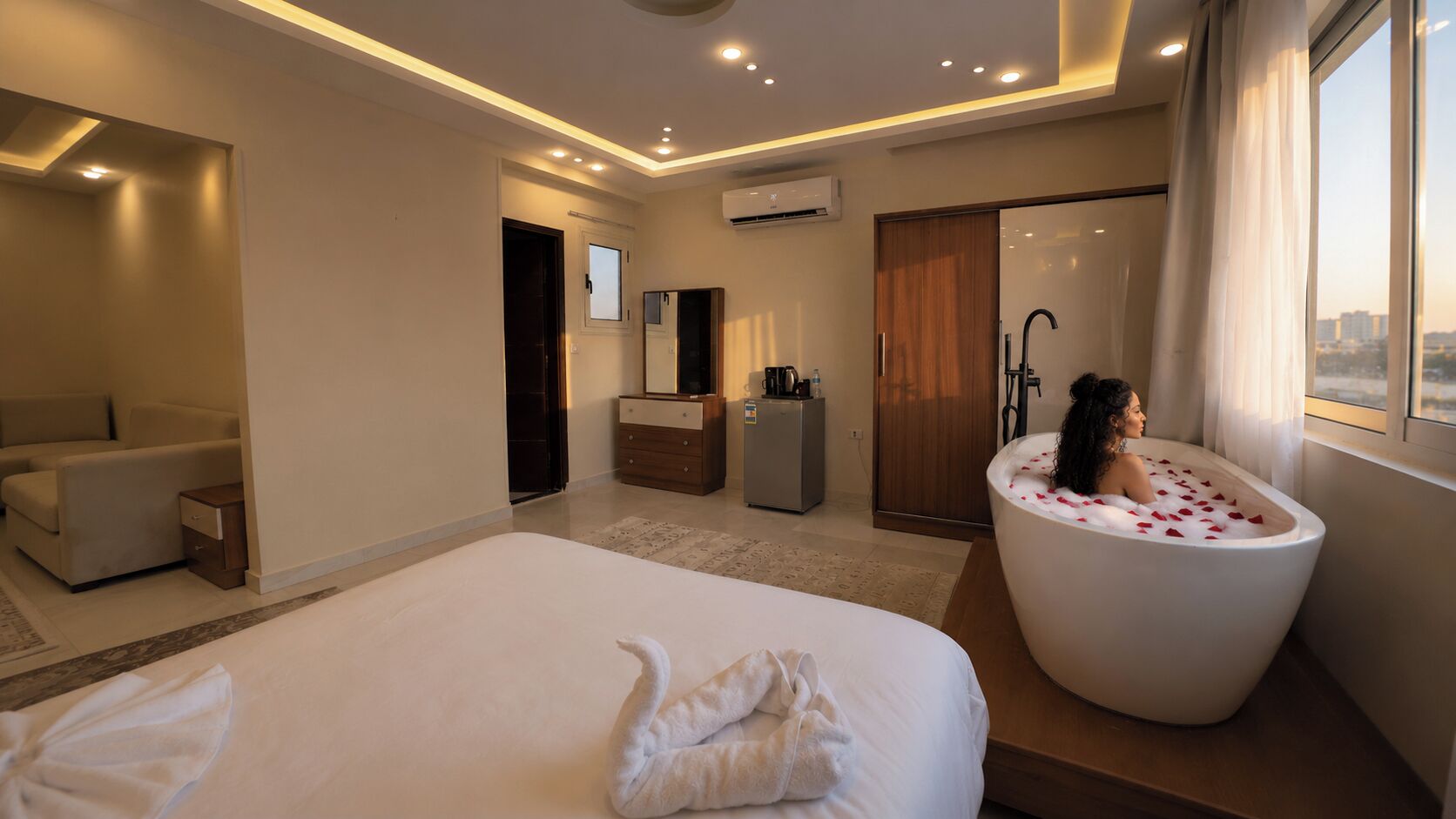 A hotel room with a large bed, a sitting area, a bathroom with a bathtub, and a view of the city.