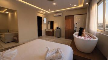 A hotel room with a large bed, a sitting area, a bathroom with a bathtub, and a view of the city.