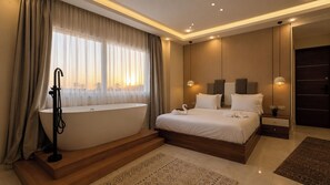 Un dormitorio moderno con una bañera grande, una cama con ropa blanca y vistas de la ciudad a través de cortinas translúcidas.