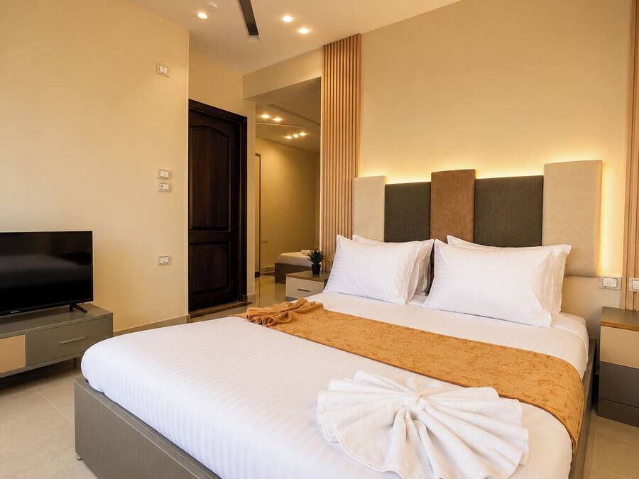 A modern hotel room with a flat-screen TV, a bed with white linens and a brown throw, a nightstand with a lamp, and a wooden door.