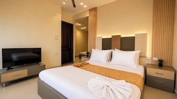 Una habitación de hotel moderna con un televisor de pantalla plana, una cama con ropa blanca y un plaid marrón, una mesita de noche con una lámpara y una puerta de madera.