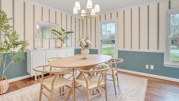 A dining room with a round table, six chairs, a chandelier, a mirror, and a view of the outdoors.