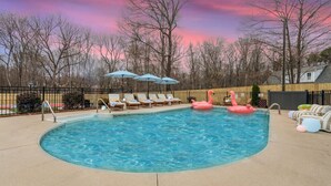 A swimming pool with lounge chairs and umbrellas, surrounded by a fence and trees. - Greensboro Getaway Villa (Greensboro)